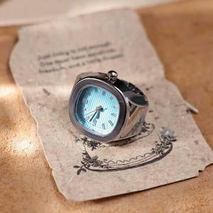 Puede incluir: Un reloj de anillo plateado con esfera cuadrada y esfera azul claro. El reloj tiene n&uacute;meros romanos y agujas negras. El anillo est&aacute; sobre un trozo de papel envejecido con texto impreso y dise&ntilde;os florales.