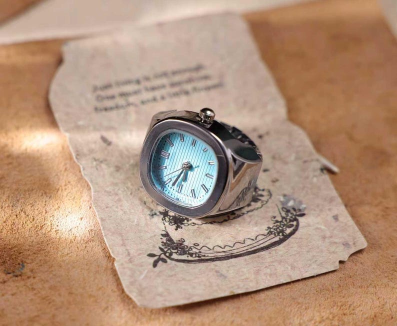 Puede incluir: Un reloj de anillo plateado con esfera cuadrada y esfera azul claro. El reloj tiene n&uacute;meros romanos y agujas negras. El anillo est&aacute; sobre un trozo de papel envejecido con texto impreso y dise&ntilde;os florales.
