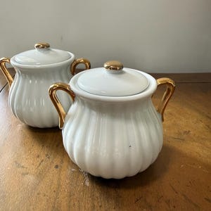 May include: Two white ceramic sugar bowls with gold-colored handles and lid accents. The bowls have a ribbed design and are set on a wooden surface. The lids have a gold knob.