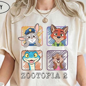 May include: Ivory-coloured t-shirt with four cartoon character portraits from Zootopia in coloured squares. The text "ZOOTOPIA 2" is printed below. The shirt is short-sleeved with a crew neck.