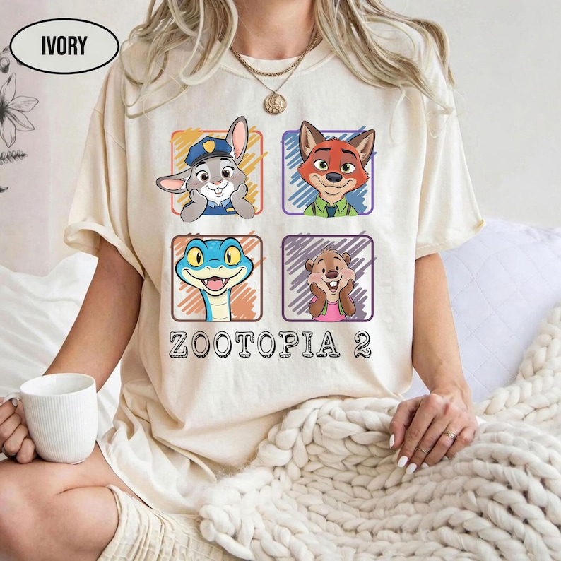 May include: Ivory-coloured t-shirt with four cartoon character portraits from Zootopia in coloured squares. The text "ZOOTOPIA 2" is printed below. The shirt is short-sleeved with a crew neck.