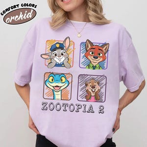May include: A lilac t-shirt with characters from Zootopia 2. The shirt displays four square graphics with cartoon images of a rabbit, fox, snake, and beaver. The text "ZOOTOPIA 2" is printed below the graphics. The shirt also has the text "COMFORT COLORS orchid".