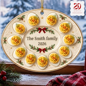 May include: An oval acrylic ornament featuring a cream-colored platter design with ten deviled eggs arranged around a central oval. The center text reads The Smith family 2026, framed by green pine branches and red bows. Gold stars and pinecones decorate the border.