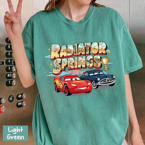 May include: Light green t-shirt with a graphic of "Radiator Springs" and images of Lightning McQueen and a blue car. The design includes a trophy and stars. The shirt is a casual, comfortable style.