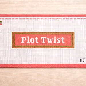 May include: A needlepoint canvas with a white background and a coral border. The phrase "Plot Twist" is stitched in white letters on a coral rectangle with a brown border. The initials "HJ" are in the bottom right corner.