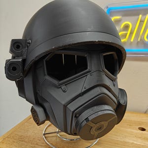 Può includere: Un casco grigio scuro futuristico con più lenti e attacchi circolari. Il casco ha una parte superiore arrotondata e una maschera facciale dettagliata con viti e prese d'aria visibili. È esposto su un supporto metallico.