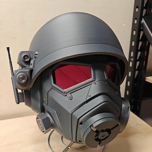 May include: A matte dark grey tactical helmet featuring two distinct red-tinted hexagonal eye lenses, a prominent central respirator filter housing, and a side-mounted communication antenna with two small circular optical sensors attached.