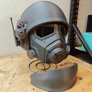 May include: A gray, futuristic-style helmet with a visor, respirator, and communication device. The helmet is displayed on a metal stand. The design includes a rounded top, angular face mask, and various mechanical details, suggesting a sci-fi or cosplay theme.