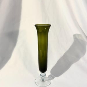 May include: A tall, olive green glass vase with a flared top and a clear, round base. The vase has a slender, cylindrical body and a decorative stem. The vase is approximately 12 inches tall, perfect for displaying flowers.