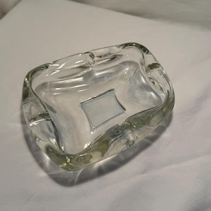 Puede incluir: Un cenicero de vidrio transparente de forma cuadrada con lados curvos. El cenicero tiene una pequeña etiqueta rectangular en el centro. Está diseñado con cuatro puntos de apoyo para cigarrillos.