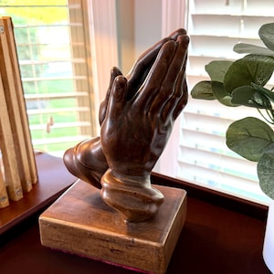 May include: A wooden sculpture of two hands clasped in prayer. The hands are a dark brown color and rest on a square wooden base. The base has a pink fabric lining the bottom edge. The sculpture is a religious home decor item.
