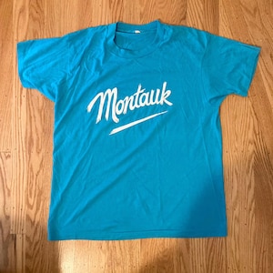 May include: A turquoise blue t-shirt with the word "Montauk" in white script across the chest. A white horizontal line is below the word. The shirt is made of a soft material and has short sleeves.