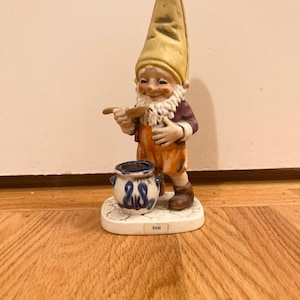 May include: A ceramic gnome figurine with a tall yellow hat, white beard, and orange overalls. The gnome holds a wooden spoon and stands next to a small blue and white pot. The base of the figurine reads "SAM".