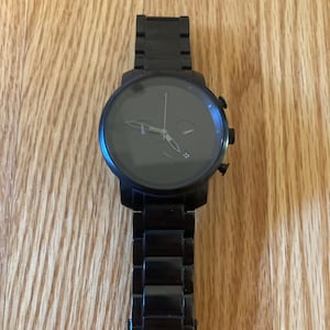 May include: A black wristwatch with a round face and a black metal strap. The watch has a dark grey face with silver hands and markers. The watch is on a wooden surface.