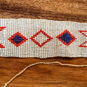 May include: A beaded bracelet with a white background and geometric patterns in red and blue. The bracelet is rectangular with white buttons on each end. The bracelet is on a wooden surface.