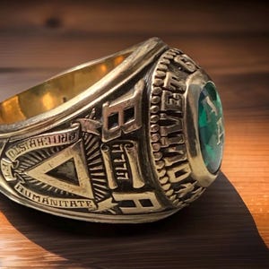May include: A gold-colored class ring with a large, emerald-green gemstone. The ring features raised lettering and symbols, including the words "PRO CHRISTO HUMANITATE" and the number "1977". The ring is likely a graduation or commemorative piece.