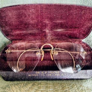May include: Vintage rimless eyeglasses with gold-tone metal frames and clear lenses, resting inside a dark purple velvet-lined case. The case is open, revealing the glasses. Text on the case reads "DR R. P. HIGGINS 20 COURT ST., CORTLAND, N.Y."
