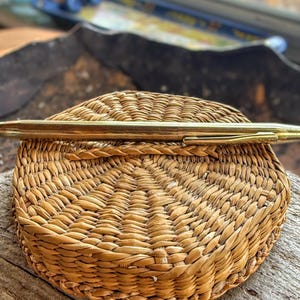 Könnte beinhalten: Ein goldfarbener Stift liegt auf einem geflochtenen Korb. Der Stift hat eine glatte, metallische Oberfläche mit einer schwarzen Spitze. Der Korb besteht aus naturfarbenem, gewebtem Material, und der Hintergrund ist unscharf.