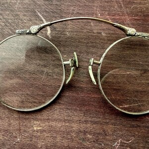 Könnte beinhalten: Antike runde Brille mit silberfarbenem Metallrahmen und klaren Gläsern. Die Brille hat dekorative Details an den Bügeln und am Steg und liegt auf einer dunklen Holzoberfläche.