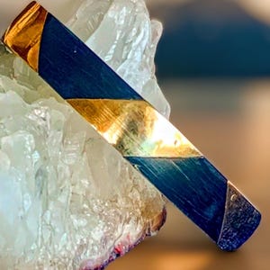 Può includere: Una barra rettangolare blu e oro con accenti dorati angolati. La barra è posta su uno sfondo di formazioni cristalline chiare. Le sezioni dorate hanno una lucentezza metallica riflettente, in contrasto con le sezioni blu più scure.