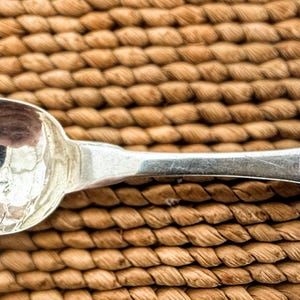 May include: A silver spoon with a rounded bowl and a rectangular handle. The handle has engraved initials. The spoon is set against a woven, brown background.