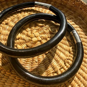 Puede incluir: Dos collares circulares negros con detalles metálicos plateados. Los collares descansan sobre una cesta tejida de color marrón claro. Los collares tienen un acabado liso y brillante y parecen estar hechos de un material sólido.