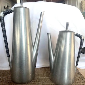 Puede incluir: Dos cafeteras de metal cepillado con boquillas largas y asas negras. Las jarras tienen un diseño alto y delgado con tapa y un pequeño asa en la parte superior. Las asas son negras y están envueltas en un material texturizado.