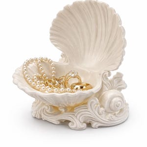 May include: A cream-coloured ceramic seashell-shaped trinket dish featuring an open clam shell design with ornate scrollwork at the base. Inside the dish rests a strand of white pearls, a gold ring with a clear stone, and a plain gold band.
