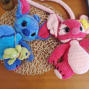 May include: Two plush, handmade crochet dolls. One is blue with pink ears and a small green doll attached. The other is pink with white accents. Both dolls are lying on a decorative, woven mat on a wooden surface.