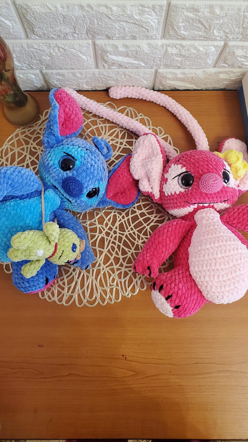 May include: Two plush, handmade crochet dolls. One is blue with pink ears and a small green doll attached. The other is pink with white accents. Both dolls are lying on a decorative, woven mat on a wooden surface.