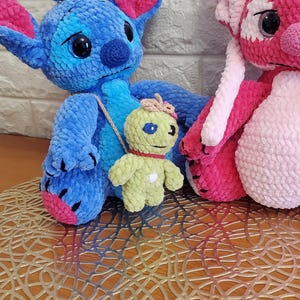 May include: Crocheted plush toys of cartoon characters, including a blue and pink creature, a pink and white creature, and a small green figure with a red collar. The toys are on a decorative gold-colored mat.