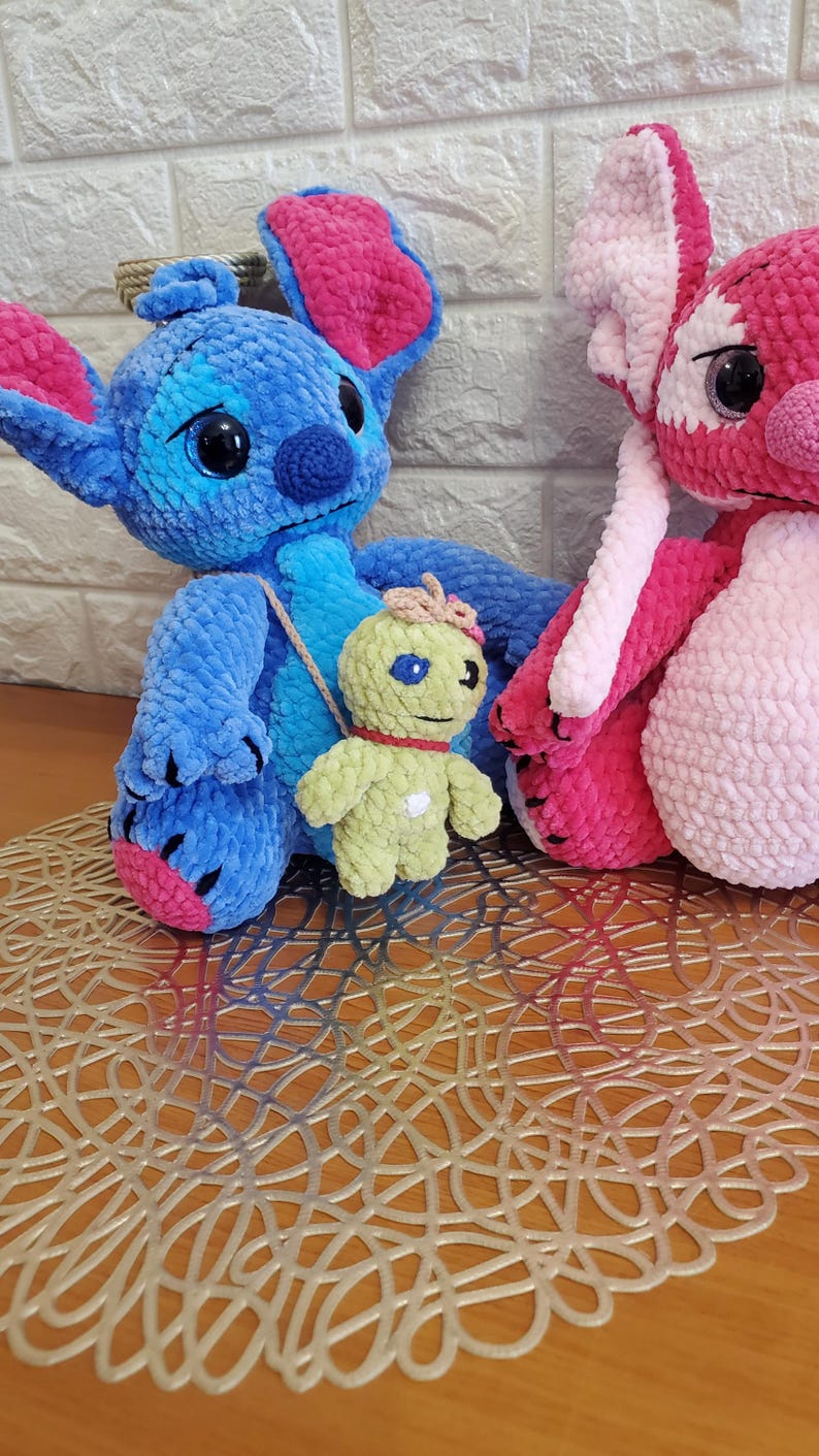 May include: Crocheted plush toys of cartoon characters, including a blue and pink creature, a pink and white creature, and a small green figure with a red collar. The toys are on a decorative gold-colored mat.
