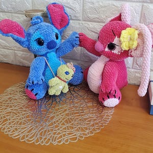 May include: Two handmade crochet plush toys. One is blue with large ears and a small green turtle-like companion. The other is pink with long ears and a yellow flower. Both are sitting on a decorative mat.