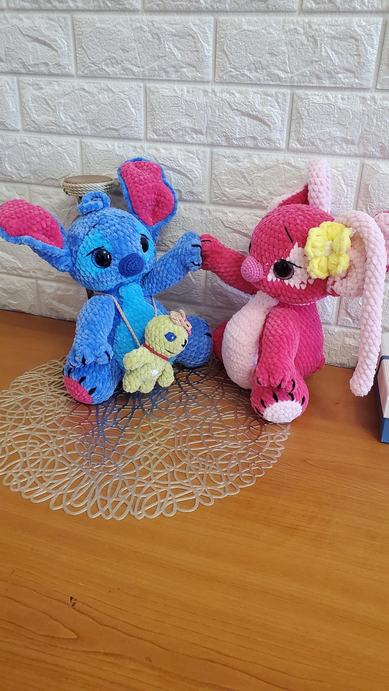 May include: Two handmade crochet plush toys. One is blue with large ears and a small green turtle-like companion. The other is pink with long ears and a yellow flower. Both are sitting on a decorative mat.