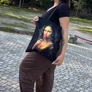 May include: Black tote bag featuring a parody of the Mona Lisa holding a lollipop. The bag is held by a person wearing a black top and brown pants. The background includes trees and a stone walkway.