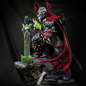 May include: A detailed action figure of Spawn, a comic book character, stands on a rocky base. The figure is clad in a dark, textured suit with a red cape and spiked gauntlets. He holds a glowing green sword, with a chain detail.