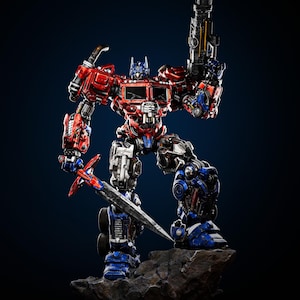 May include: A detailed statue of Optimus Prime, a red, blue, and silver robot, holding a large gun and a sword. The figure is posed on a rocky base against a dark blue background. The robot's design features intricate mechanical details.