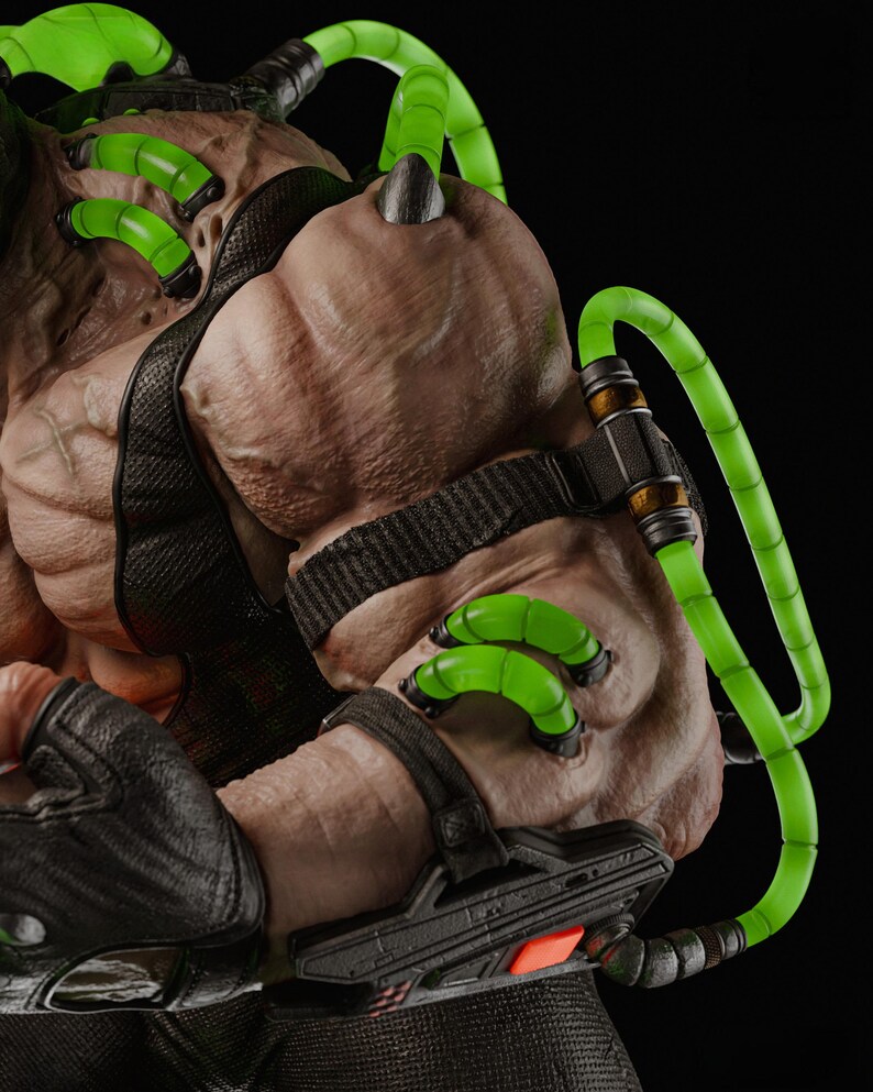 Op de afbeelding: Close-up van een gespierde figuur met een getextureerde huid. De figuur heeft zwarte banden en buizen gevuld met felgroene vloeistof. De figuur draagt zwarte handschoenen en heeft een mechanisch apparaat op de arm. De achtergrond is zwart.