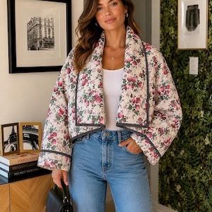 Puede incluir: Chaqueta acolchada con un estampado floral de fresas rosas sobre fondo crema. La chaqueta tiene ribetes negros y se combina con vaqueros azul claro. Se sostiene un bolso negro. Una opción de abrigo elegante.