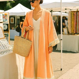 Può includere: Una donna indossa un lungo abito color pesca aperto con frange, un vestito color crema e una borsa di paglia. Indossa occhiali da sole e sandali. La scena è un mercato all'aperto con tende bianche ed espositori.