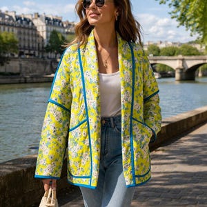 May include: A yellow floral print jacket with blue trim, worn with a white top and blue jeans. The jacket has a quilted design. The woman is wearing sunglasses and carrying a small woven bag. The background shows a river and buildings.