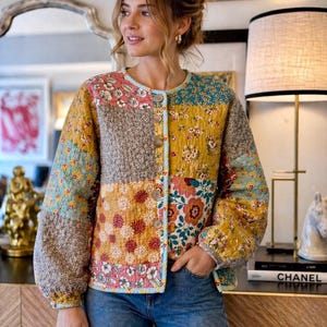 May include: A multi-coloured patchwork jacket with a round neck and button fastening. The jacket combines floral and patterned fabrics in yellow, blue, pink, and brown. The sleeves are long and slightly puffed, worn with blue jeans.