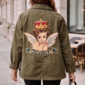 Könnte beinhalten: Olivgrüne Jacke mit großem, aufgesticktem Aufnäher auf dem Rücken, der einen Cherub mit Krone und Flügeln sowie den Text "DGDEVOTION" zeigt. Die Jacke hat einen Kragen und geknöpfte Manschetten. Die Person trägt eine schwarze Hose und eine schwarze Handtasche.