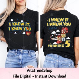 Pu&ograve; includere: Una t-shirt nera con un design grafico. Il davanti mostra il testo "I KNEW IT, I KNEW YOU" con un'icona a forma di cuore. Il retro mostra il testo "I KNEW IT I KNEW YOU we're TURNING 5" con illustrazioni di cappello da cowboy, blocchi e stivali.