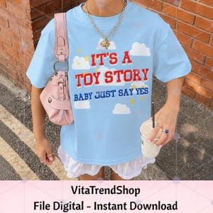 K&ouml;nnte beinhalten: Ein hellblaues T-Shirt mit dem roten Text IT'S A TOY STORY und dem blauen Text BABY JUST SAY YES, verziert mit wei&szlig;en Wolken- und gelben Sternengrafiken. Das T-Shirt wird mit einer rosa Umh&auml;ngetasche und einem wei&szlig;en R&uuml;schenrock kombiniert.