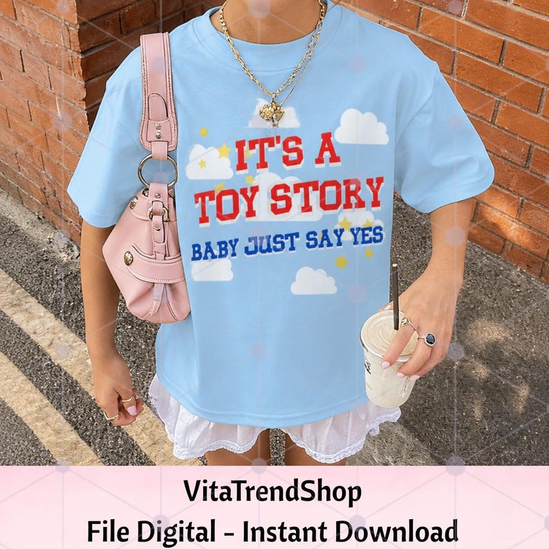 K&ouml;nnte beinhalten: Ein hellblaues T-Shirt mit dem roten Text IT'S A TOY STORY und dem blauen Text BABY JUST SAY YES, verziert mit wei&szlig;en Wolken- und gelben Sternengrafiken. Das T-Shirt wird mit einer rosa Umh&auml;ngetasche und einem wei&szlig;en R&uuml;schenrock kombiniert.