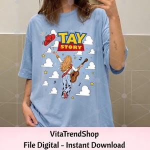 May include: A light blue t-shirt featuring a graphic of a person with long blonde hair playing an acoustic guitar, wearing cow-print pants and a red cowboy hat. The text TAY STORY is printed above, surrounded by white clouds and yellow stars.