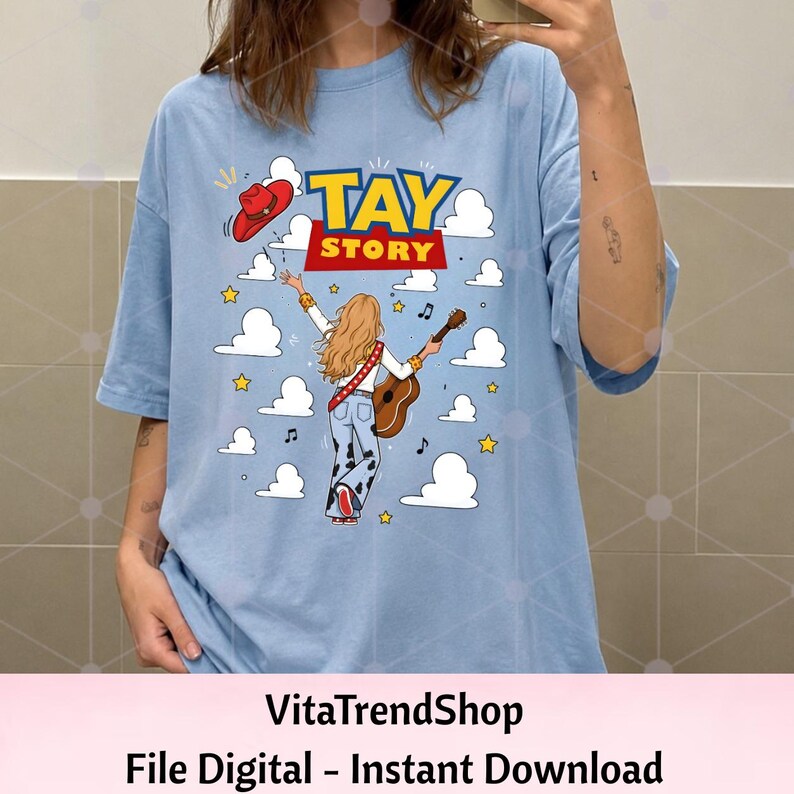 May include: A light blue t-shirt featuring a graphic of a person with long blonde hair playing an acoustic guitar, wearing cow-print pants and a red cowboy hat. The text TAY STORY is printed above, surrounded by white clouds and yellow stars.