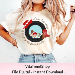 May include: A cream-colored t-shirt featuring a graphic of a black tire framing a scene with cartoon characters, a red cowboy hat, and a red cowboy boot. The text reads, Love has ways of bringing things back to life, with the name Taylor on the boot.