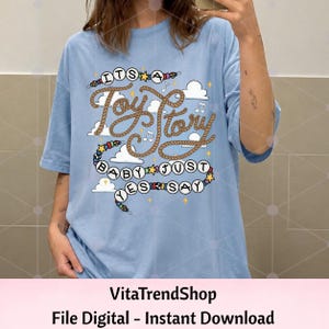 May include: A light blue t-shirt featuring a graphic print with the text "ITS A Toy Story BABY JUST YES SAY". The design includes brown rope-style lettering, white clouds, musical notes, yellow stars, and a string of colorful beads.